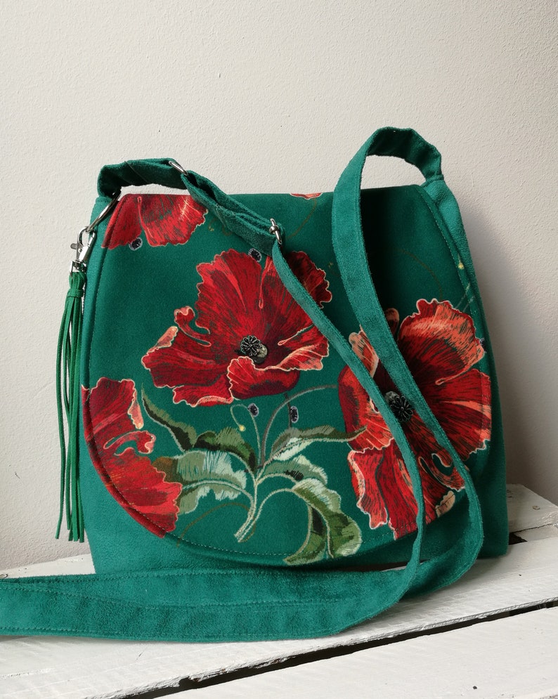 May include: A green suede shoulder bag with a floral print featuring red poppies. The bag has a long strap and a tassel.