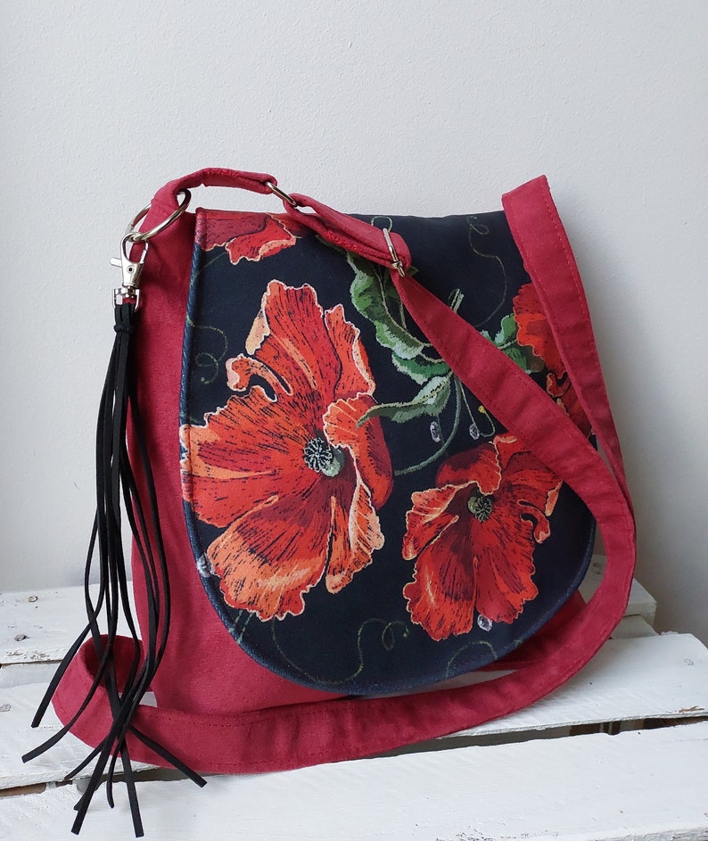 May include: A red crossbody bag with a black floral print featuring red poppies. The bag has a long strap and a black tassel.