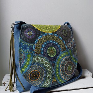 May include: A blue denim shoulder bag with a flap featuring a colorful mandala pattern in shades of blue, green, yellow, and purple. The bag has a long strap and a decorative olive green tassel.
