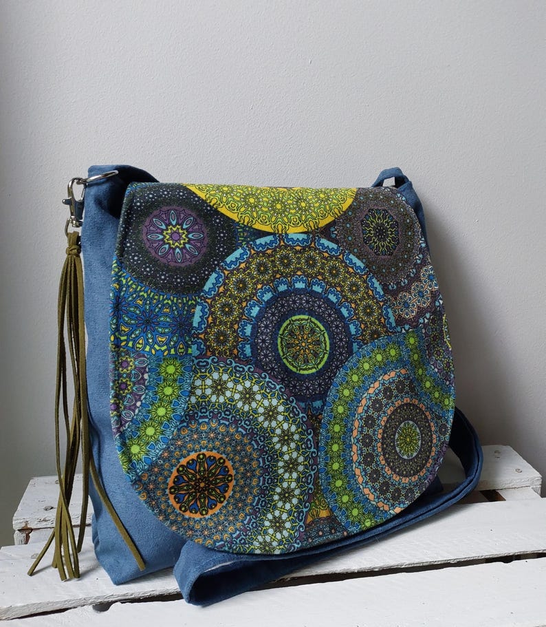 May include: A blue denim shoulder bag with a flap featuring a colorful mandala pattern in shades of blue, green, yellow, and purple. The bag has a long strap and a decorative olive green tassel.
