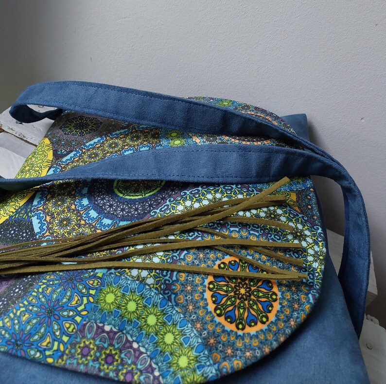 May include: A blue denim shoulder bag with a colorful, patterned flap. The flap features a vibrant mandala design in shades of blue, green, yellow, and orange. Olive green fringe hangs from the flap. The bag has a blue denim strap.