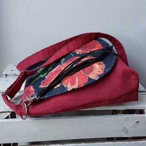 May include: A red crossbody bag with a flap closure. The flap is made of a dark fabric with a floral print of red poppies on a black background. The bag has a black tassel.