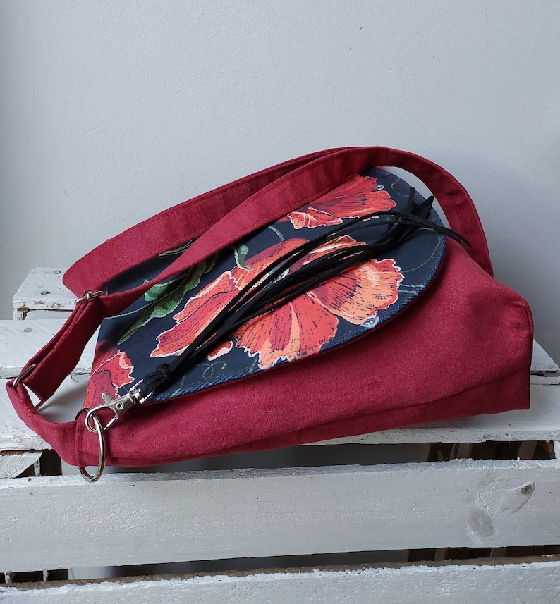 May include: A red crossbody bag with a flap closure. The flap is made of a dark fabric with a floral print of red poppies on a black background. The bag has a black tassel.