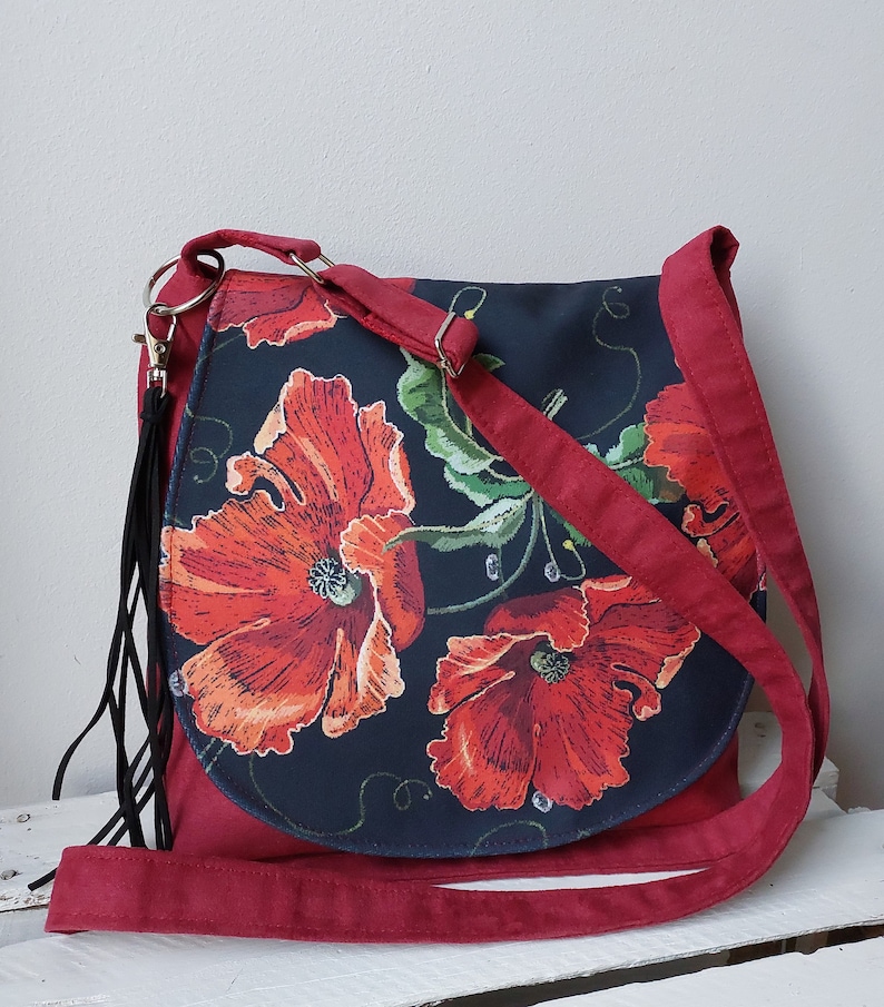 May include: A red suede crossbody bag with a black fabric flap featuring a floral design of red poppies on a black background.