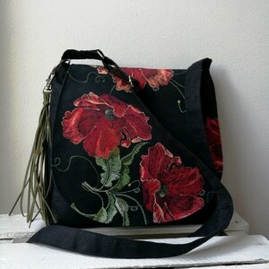 May include: A black canvas shoulder bag with a long strap and a green tassel. The bag features a floral design with red poppies and green leaves.