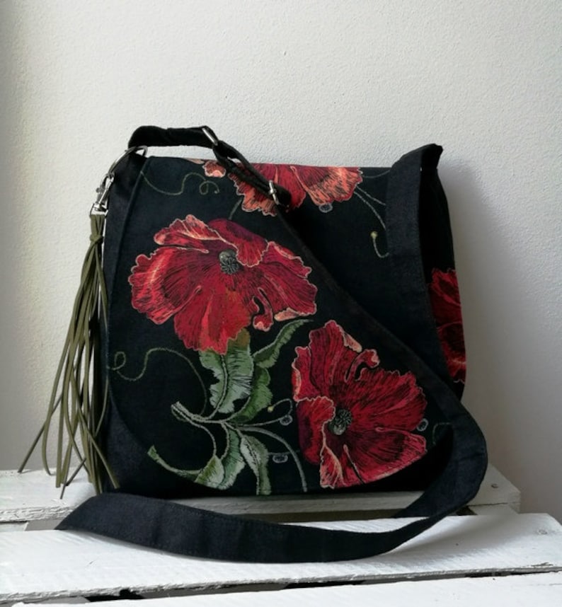 May include: A black canvas shoulder bag with a long strap and a green tassel. The bag features a floral design with red poppies and green leaves.