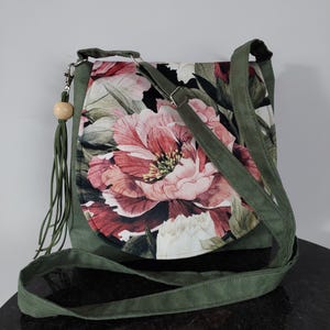 May include: A green canvas crossbody bag with a floral print featuring large pink flowers. The bag has a long adjustable strap and a wooden bead tassel.
