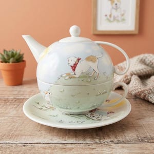 May include: A ceramic tea-for-one set comprising a teapot that stacks onto a teacup and saucer. The set is decorated with illustrations of white dogs with brown patches wearing red bandanas, set against a soft blue and green landscape background.