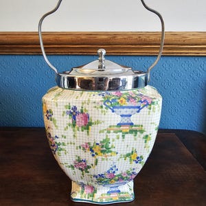 May include: A vintage ceramic biscuit jar with a floral pattern in pink, yellow, and blue. It has a silver-toned lid and handle. The jar is on a dark wooden surface.