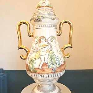 May include: A white ceramic vase with gold handles and accents. The vase features a painted scene of cherubs and foliage. The lid is decorated with a floral design. The vase is displayed on a mirrored surface.
