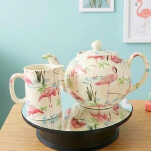 May include: A cream-coloured ceramic teapot and matching creamer pitcher featuring a repeating pattern of pink flamingos, green palm leaves, and blue water accents. Both pieces have a glossy finish and are displayed on a round mirrored surface.