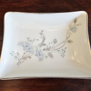 May include: A rectangular white porcelain dish with rounded corners and a gold trim. The dish is decorated with a floral pattern in shades of blue, grey, and green. The flowers and leaves are delicately painted.