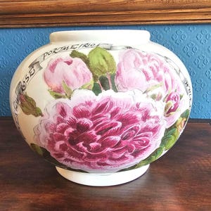 May include: A white ceramic vase with a round shape, featuring a large pink peony flower design. The vase has a wide opening and a base. The words "Rose Pormetrio" are written on a banner around the vase.