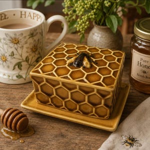 May include: A rectangular ceramic butter dish with a raised honeycomb pattern in shades of brown and yellow, topped with a sculpted bee figure. The dish sits on a matching rectangular base, featuring a glossy finish and a detailed hexagonal design.