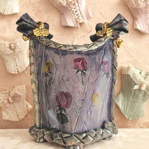 May include: A square ceramic container with a textured lavender surface featuring painted rose motifs. The top and bottom edges are framed with a braided metallic border. Two dark fabric bows with gold-toned accents are attached to the top corners.