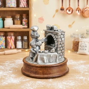 May include: A silver-toned pewter figurine depicting two bears dressed as bakers, one placing a tray into a brick oven and the other standing at a table with dough, all mounted on a circular wooden base with a dusting of flour on the surface.