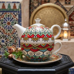 May include: A ceramic tea set for one, consisting of a teapot that stacks onto a matching teacup, both resting on a saucer. The set features a vibrant, repeating pattern in red, teal, yellow, and black, with gold-toned trim on the lid and rims.