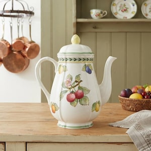 May include: A white ceramic teapot with a fluted body and a matching lid featuring a yellow knob. The teapot is decorated with colourful illustrations of cherries, plums, and pears, accented by a green leaf border pattern around the top and base.