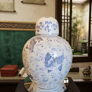 May include: A white ceramic ginger jar with a matching lid, featuring blue hand-painted illustrations of koi fish swimming among aquatic plants and pine needles. The rounded vessel has a smooth, glossy finish and a classic bulbous silhouette.