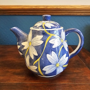 May include: A blue ceramic teapot with a lid, handle, and spout. The teapot is decorated with white daisy-like flowers with yellow centres and stems. The background is a dark wood surface.