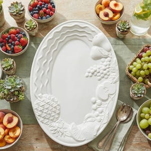 May include: An oval white ceramic serving platter featuring embossed fruit designs including a pineapple, grapes, and citrus slices along the rim, with a wavy textured border pattern on the left side and a smooth centre surface for food presentation.