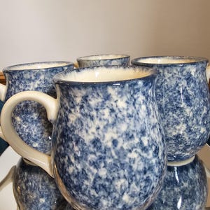 May include: Four blue and white speckled ceramic mugs with white handles and rims. The mugs are arranged on a reflective surface, showcasing their speckled design and classic shape. The mugs are a classic kitchenware item.