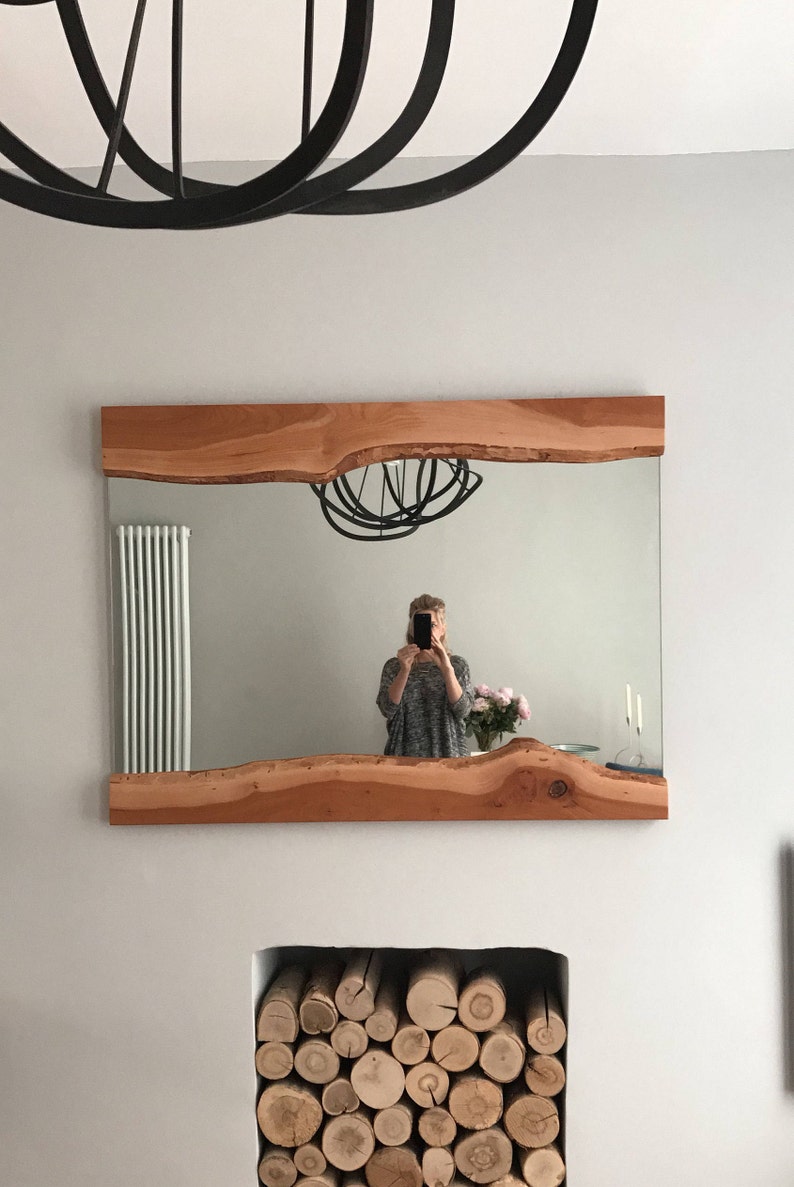 Puede incluir: Un espejo con marco de madera y borde natural, que refleja a una persona tomando una selfie y una l&aacute;mpara de ara&ntilde;a de metal negro. El espejo est&aacute; montado en una pared blanca sobre una chimenea con troncos apilados.
