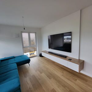 May include: A living room featuring a teal sectional sofa, light wood flooring, and a wall-mounted entertainment unit with white cabinet doors and a wood frame. A large flat-screen television is mounted above the unit with a black soundbar below.