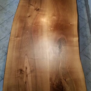 May include: A large, natural wood slab table top with a live edge. The wood has a warm, honey brown color and a smooth, polished finish. The grain pattern is visible and adds to the rustic charm of the table.