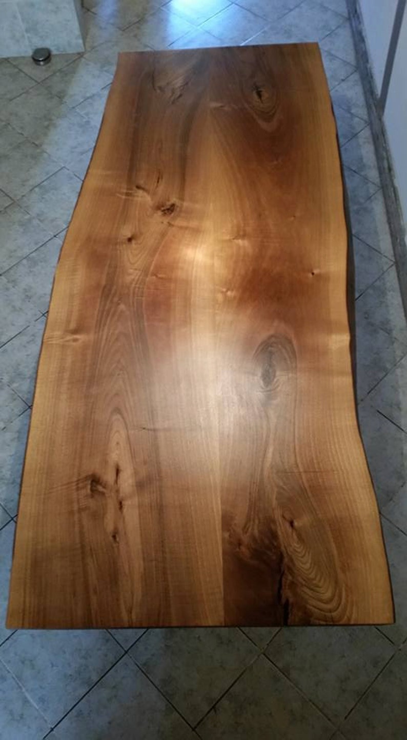 May include: A large, natural wood slab table top with a live edge. The wood has a warm, honey brown color and a smooth, polished finish. The grain pattern is visible and adds to the rustic charm of the table.