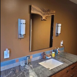 May include: A bathroom vanity with a large mirror framed by a natural wood slab. The countertop is gray and white veined granite, with a white sink. Two sconces flank the mirror, and the cabinetry is a warm wood tone.