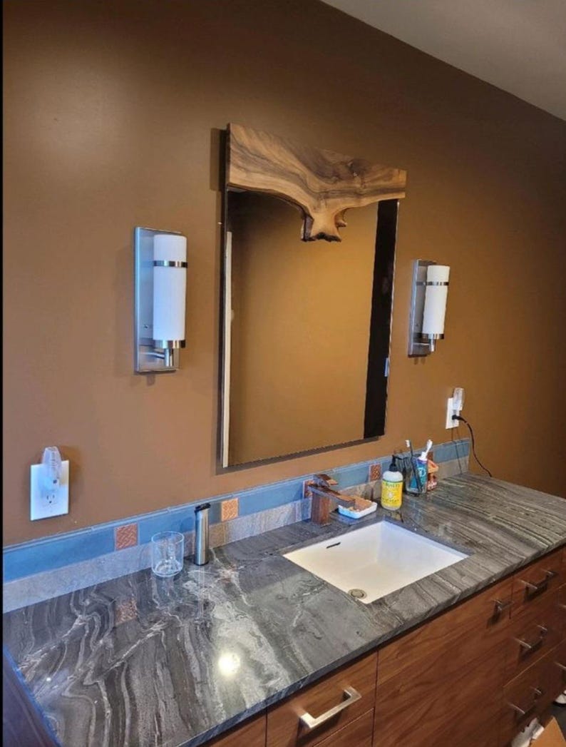 May include: A bathroom vanity with a large mirror framed by a natural wood slab. The countertop is gray and white veined granite, with a white sink. Two sconces flank the mirror, and the cabinetry is a warm wood tone.