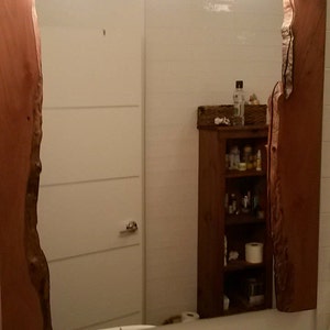 Puede incluir: Un espejo de ba&ntilde;o con un marco de madera. El marco est&aacute; hecho de dos piezas grandes de madera con un borde natural. El espejo refleja una puerta blanca, un armario de madera con estantes y un lavabo con un grifo.
