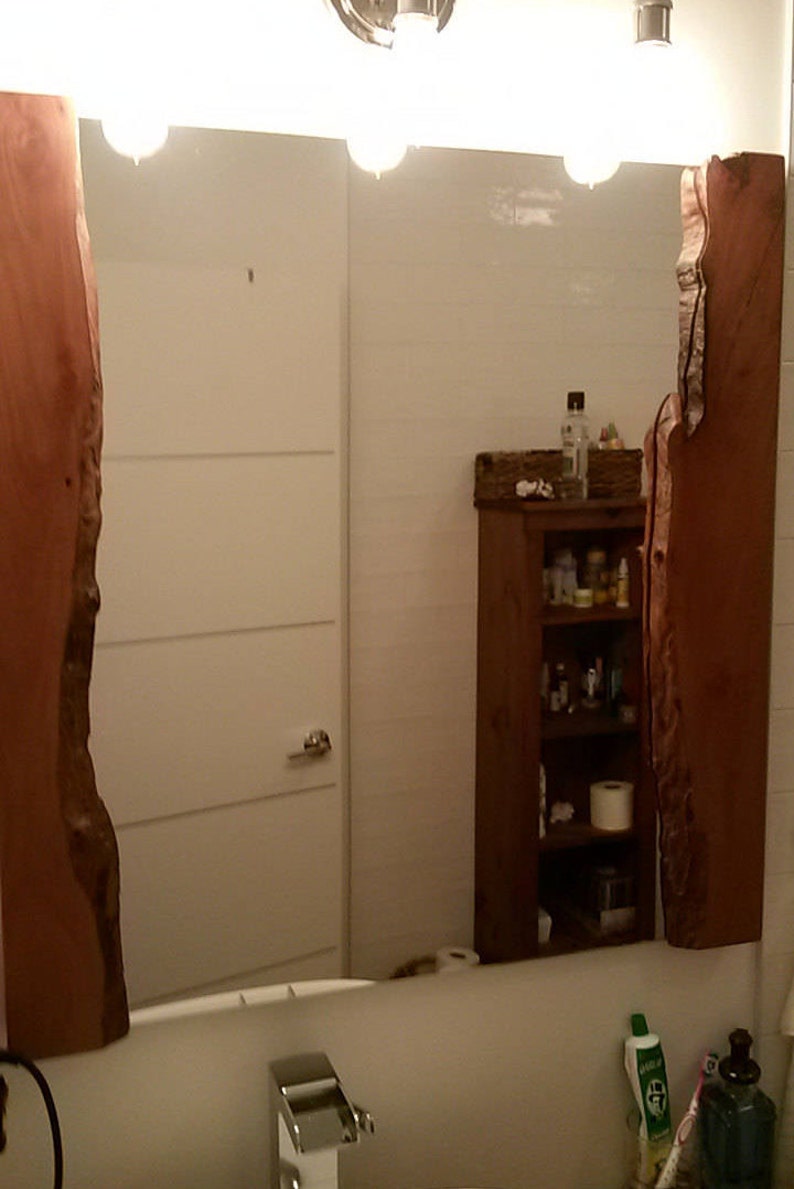 Puede incluir: Un espejo de ba&ntilde;o con un marco de madera. El marco est&aacute; hecho de dos piezas grandes de madera con un borde natural. El espejo refleja una puerta blanca, un armario de madera con estantes y un lavabo con un grifo.