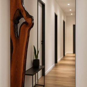 May include: A rectangular mirror with a black frame and a natural wood accent on the left side. The wood has a unique, organic shape with a dark, open knot. The mirror reflects a hallway with black doors and a wooden floor.