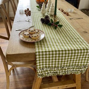 May include: A green and white gingham table runner with a ruffled edge, placed on a wooden table. The runner is decorated with three glass Christmas tree ornaments, pinecones, holly, two green taper candles, and a plate of star-shaped mince pies.