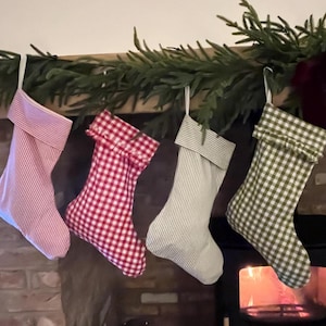 May include: Four fabric stockings hanging in a row. The first is pink and white striped, the second is red and white gingham, the third is sage green and white striped, and the fourth is sage green and white gingham. Each has a folded cuff.