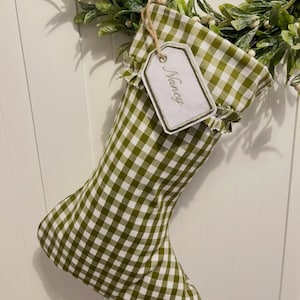 May include: A Christmas stocking featuring a green and white gingham check pattern with a ruffled cuff. A white fabric gift tag with a green border is attached to the stocking with twine, displaying the embroidered name Nancy in green script.