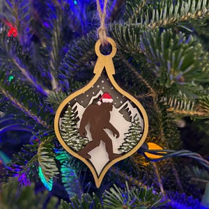 Op de afbeelding: Een houten kerstornament met een bruine Bigfoot met een kerstmuts, tegen een besneeuwde bergachtergrond. Het ornament heeft een goudkleurige rand en hangt aan een touwtje. De achtergrond is een kerstboom.