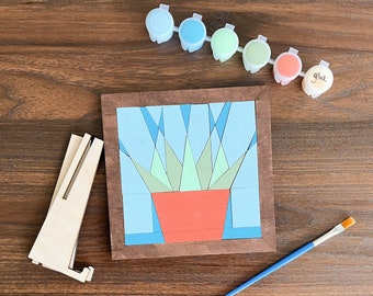 Puede incluir: Un marco de madera con un diseño geométrico de una planta en una maceta. El marco está rodeado de botes de pintura en varios tonos de azul y verde, junto con un pincel y un caballete de madera.
