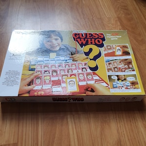 May include: A board game box for the game Guess Who? The box is red, white, and blue with a large question mark on the front. The game is described as a "killer mystery-face game".