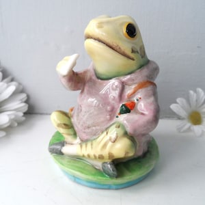 Op de afbeelding: Een keramieken kikkerfiguur met een roze jas en zittend op een waterlelieblad. De kikker heeft een gele kop, zwarte ogen en houdt een klein wit voorwerp vast. Een vis ligt aan de voeten van de kikker.