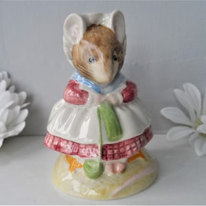 May include: A ceramic figurine of a mouse wearing a bonnet, dress, and shawl. The mouse is holding a green knitting needle and yarn. The dress has red trim and the figurine is on a yellow base.