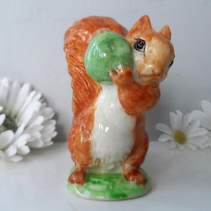 May include: A ceramic squirrel figurine with a bushy orange tail and a white belly, holding a green object. The squirrel is standing on a green base. Daisies are in the background.