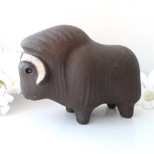 May include: A brown ceramic figurine of a musk ox with white accents on the horns. The figurine is standing on a white surface.
