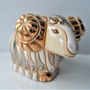 May include: A white ceramic figurine of a ram with gold and silver accents. The ram has large, curled horns and a detailed face.