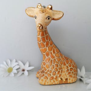 May include: A ceramic giraffe figurine with a tan and orange spotted pattern. The giraffe has gold accents on its ears, horns, and snout. White flowers are scattered around the base of the figurine. The giraffe is a decorative item.