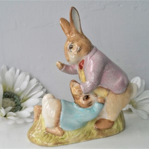 May include: A ceramic figurine of two rabbits. One rabbit, in a pink jacket and red bow tie, stands over another in a blue shirt. The base is painted to look like grass, with a yellow and green color scheme.