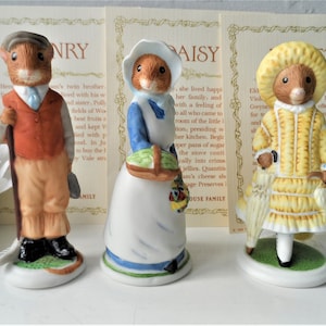 Puede incluir: Tres figuras de cerámica de ratones antropomórficos. Un ratón lleva una gorra marrón, un chaleco naranja y lleva un palo de golf. Otro ratón lleva un vestido y un gorro azules y blancos, sosteniendo una cesta. El tercer ratón lleva un vestido y un gorro amarillos, sosteniendo una sombrilla.