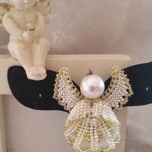 以下が含まれることがあります： 真珠の中心を持つ白と金のビーズでできた天使のオーナメント。天使は羽根と流れるようなガウンをしています。